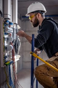 A male electrician works in a switchboard with an electrical connecting cable, connects the equipment with tools.